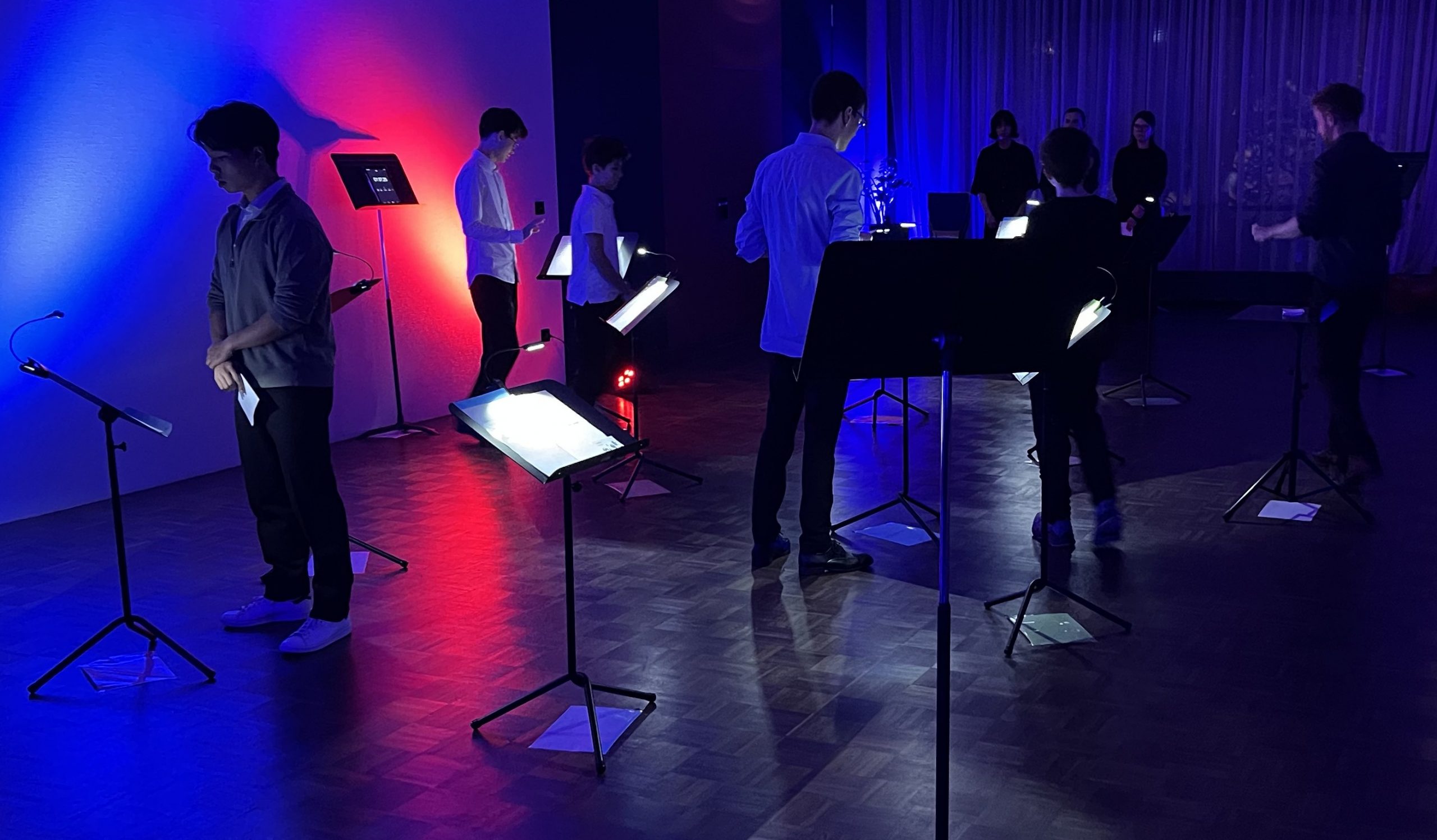 In einem düsteren, blau und rot beleuchteten Raum stehen vereinzelt schwarze Notenständer; dazwischen gehen und stehen einzelne Jugendliche