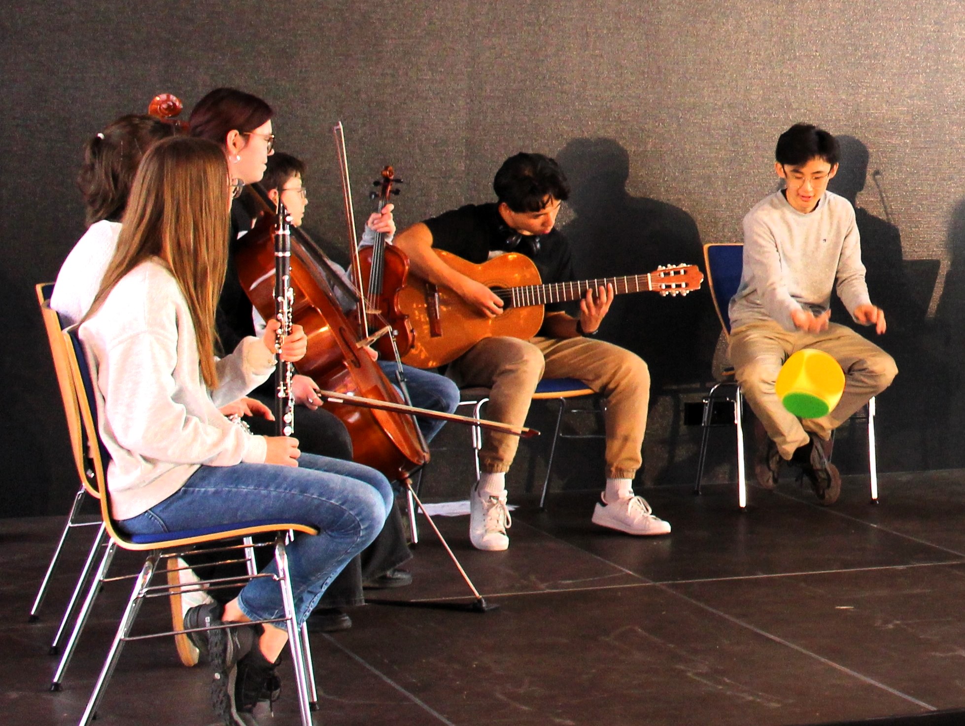 Jugendliche mit Musikinstrumenten sitzen im Halbkreis, einer von ihnen wirft einen großen Würfel mit farbigen Seiten